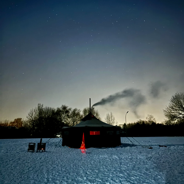 Eine Jurte steht im Schnee bei Dunkelheit. Aus dem Zelt dringt Feuerschein.